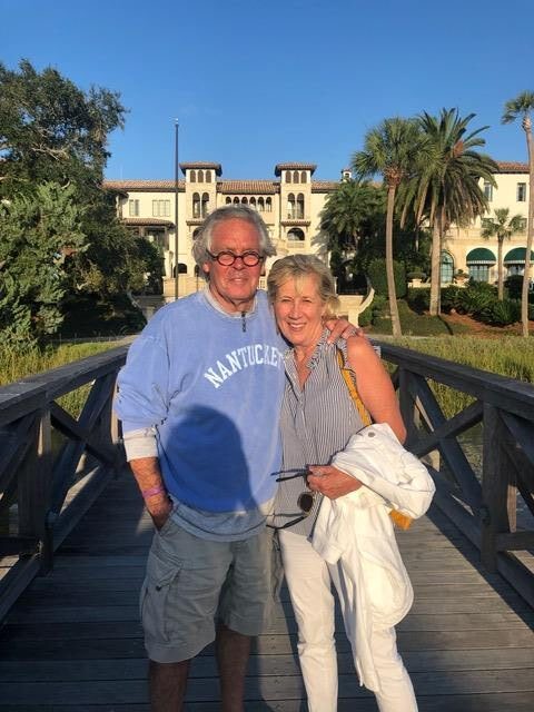 Mary Catherine, a blonde woman in middle age wearing a blue sweater, leans affectionately towards her husband, Greg, also middle aged. He has white hair and wears glasses, He is also wearing a blue sweater.